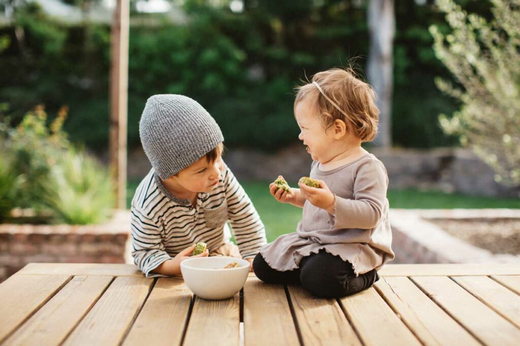 Two children sit on a deck enjoying broccoli balls from a bowl.