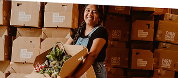 A person holds a Harvest Box full of produce with many boxes stacked high behind them.
