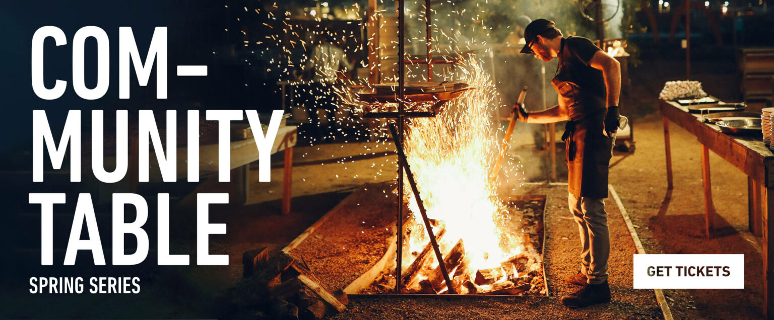 Community Table promotional banner with a chef cooking at night over an open fire