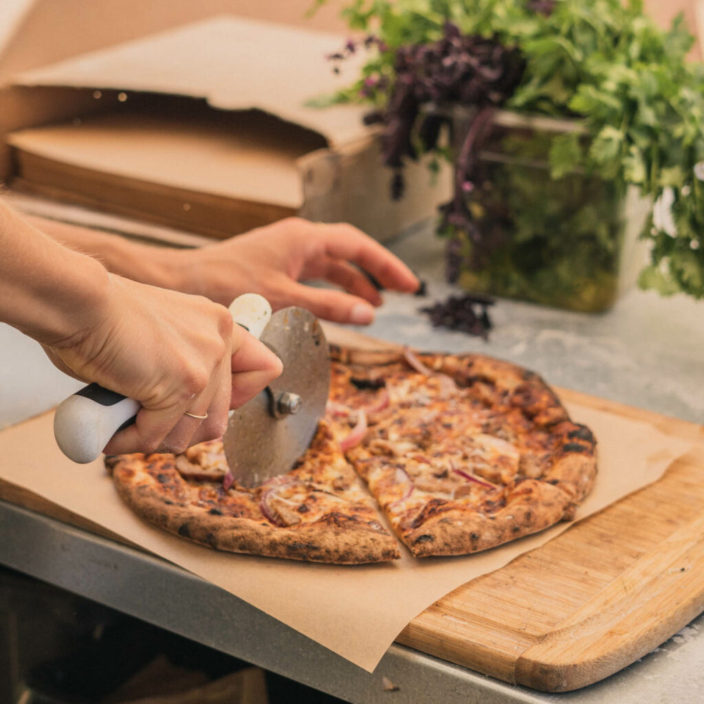 A person cuts a wood fire pizza on a wooden cutting board.