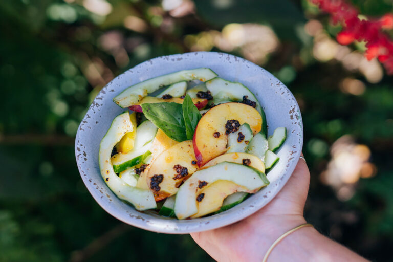 hand holding out a bowl of cucumber stone fruit salad drizzled in salsa macha