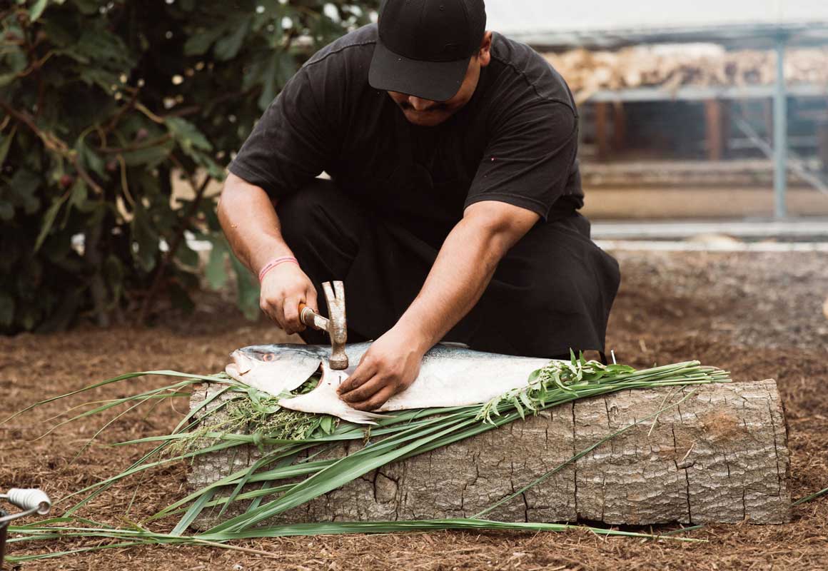 Chef prepares a large fish over leaves and a log.
