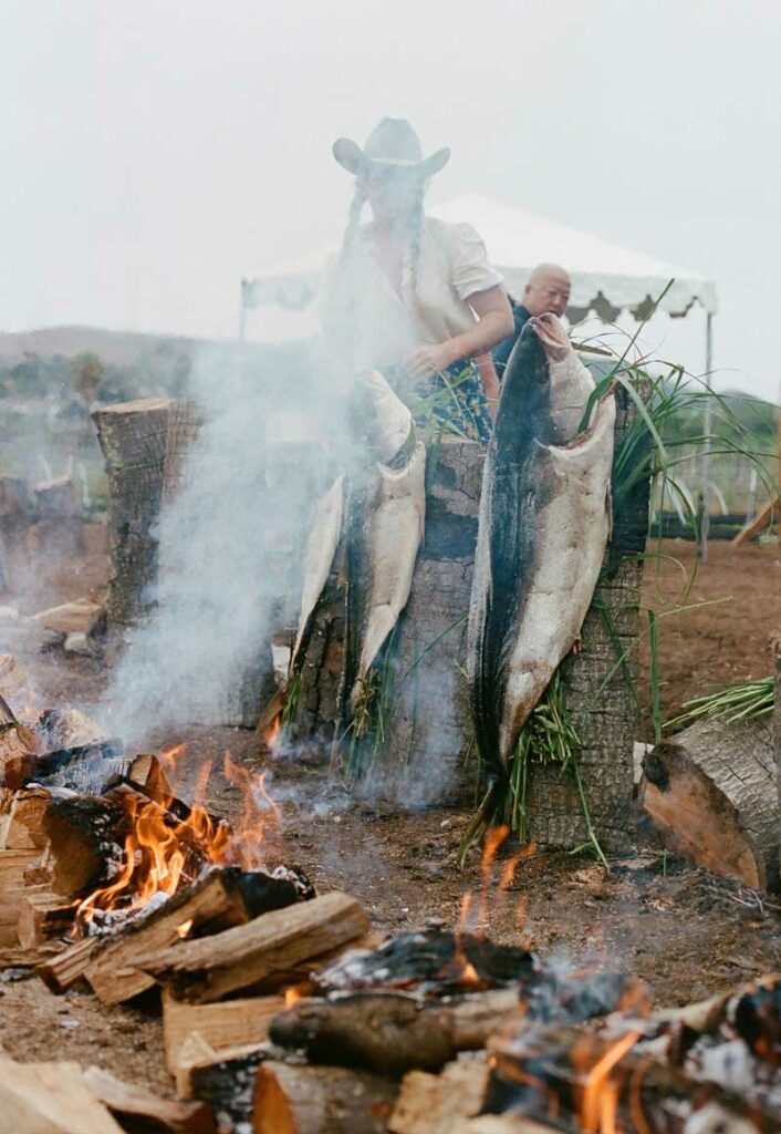 A chef is obscured behind a plume of white spoke from the open fire roasting fish.